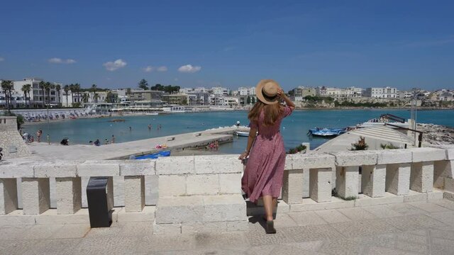 Young woman with dress and hat walking towards Otranto promenade, Salento, Italy