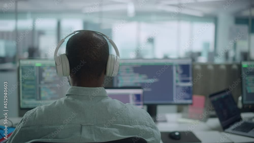 Poster Office: Professional Black IT Programmer Uses Headphones while Working on Desktop Computer. Male Website Developer, Software Engineer Developing App, Video Game. Listening to Podcast, Music. Back View