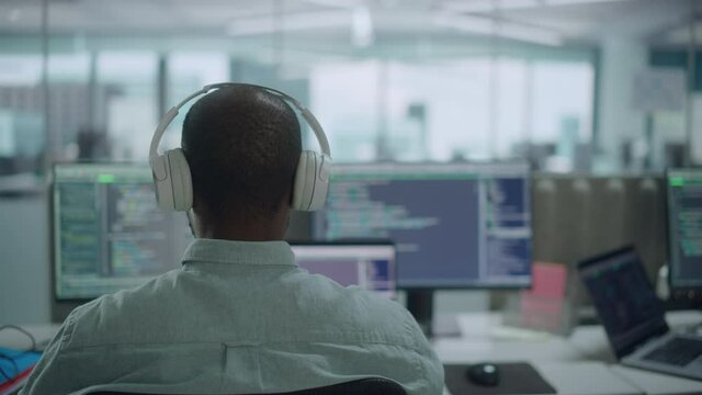 Office: Professional Black IT Programmer Uses Headphones while Working on Desktop Computer. Male Website Developer, Software Engineer Developing App, Video Game. Listening to Podcast, Music. Back View