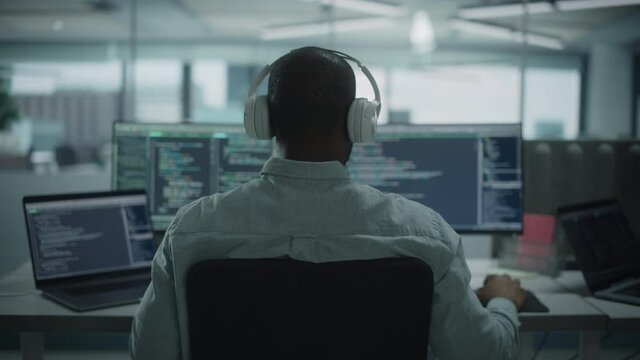 Office: Professional Black IT Programmer Uses Headphones while Working on Desktop Computer. Male Website Developer, Software Engineer Developing App, Video Game. Listening to Podcast, Music. Back View