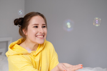 happy brunette woman in yellow hoodie among soap bubbles in bright grey room. happy people. millennial generation. fashionable teenager. trendy colours.