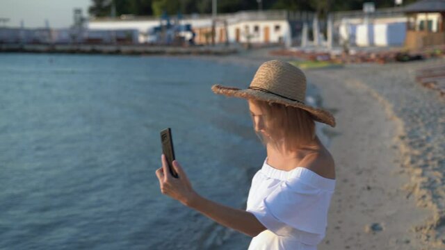 A young Caucasian woman walks near the sea or ocean early in the morning at dawn, holds a smartphone in her hands and makes a video call. 4k