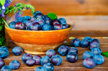A clay cup with ripe plums and individual plums lying side by side on a wooden table 