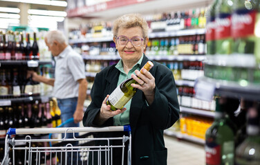 mature woman with glasses chooses bottle of wine in alcohol section of supermarket