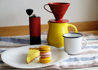 Manual brewing coffee still life in pastoral style aesthetics. Enamel mug, yellow ceramic jug, red dripper and coffee grinder. Macaroons and lemon tart on white plate