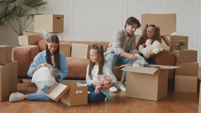 Big Moving day. Happy family of four unpacking boxes in new home, cheerful little twin sisters helping parents