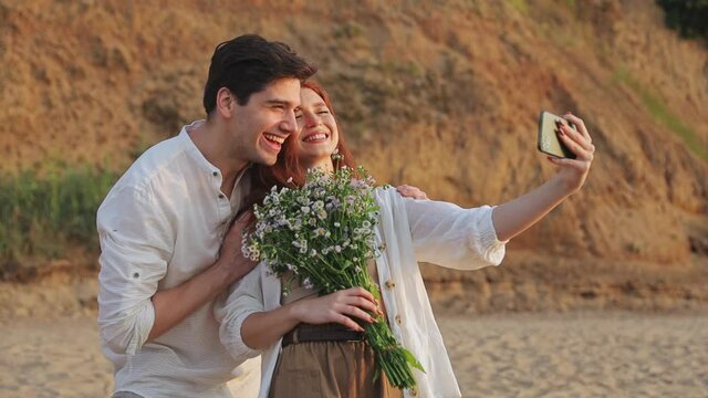 Young couple two friends family man woman 20s in white clothes hold flowers bouquet doing selfie shot on mobile cell phone at sunrise over sea beach ocean outdoor seaside in summer day sunset evening