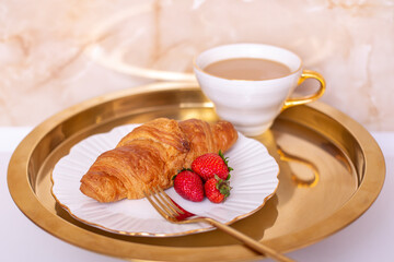 Coffee cup and sweet croissant with fresh strawberry on golden plate