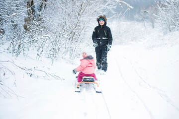 little girl of 1 year old sits on sled. brother takes sister along snow-covered road in countryside. Heavy snowfall. Bad weather in winter