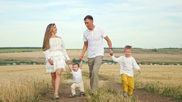 Family couple in white clothes walks joining hands with children and lifts toddler son in yellow wheat field in hot summer, against blue sky on horizon