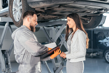 Auto Mechanic holding clipboard talking to female customer in workshop at auto repair center....