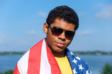 Portrait of young African American man in the sunglasses looking at camera and covered with American flag on his shoulders against blue sky 
