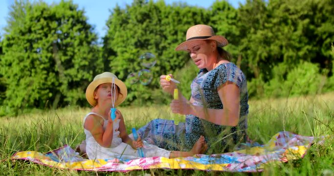 A pregnant mother and her five-year-old daughter play together, blow and catch soap bubbles while sitting in the park. Mother's Day celebration. Happy family playing in the park or forest, slow motion