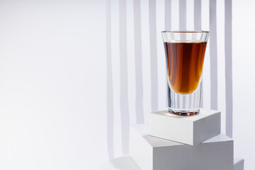 Bright amber brandy cocktail in shot glass in sunlight on white square podium as stairs with abstract stripes in soft light space in luxury minimal geometric style.