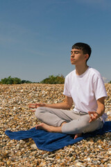 Naklejka na ściany i meble A Caucasian teenage boy meditating on a stony beach with his hands in the Shuni position