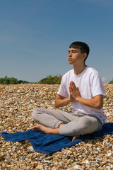 A Caucasian teenage boy meditating on a stony beach with his hands in a prayer position