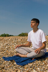 Naklejka na ściany i meble A Caucasian teenage boy meditating on a stony beach with his hands resting on his knees