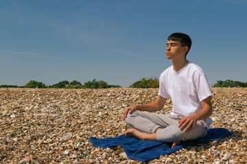 Fototapeta na wymiar A Caucasian teenage boy meditating on a stony beach with his hands resting on his knees