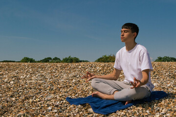 Fototapeta na wymiar A Caucasian teenage boy meditating on a stony beach with his hands in the Shuni position