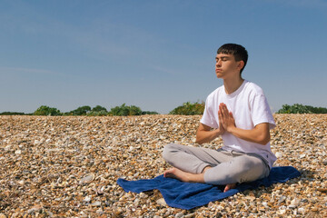 Naklejka na ściany i meble A Caucasian teenage boy meditating on a stony beach with his hands in a prayer position