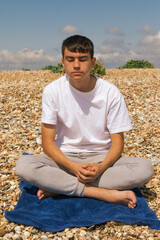 Naklejka na ściany i meble A Caucasian teenage boy meditating on a stony beach with his hands resting on his lap