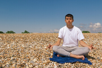 Naklejka na ściany i meble A Caucasian teenage boy meditating on a stony beach with his hands in the Shuni position