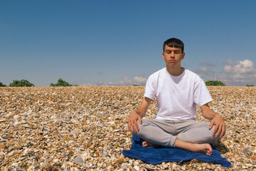 A Caucasian teenage boy meditating on a stony beach with his hands resting on his knees