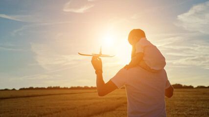 Young father carries toddler son on shoulders and toy red plane in hand and turns round flying airplane in yellow field at sunset light, sunlight