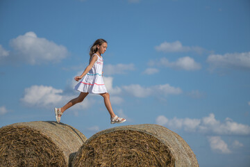 Naklejka na ściany i meble A beautiful girl plays in the field on bales of straw.