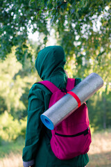 A Muslim woman in a green jacket and a blue hijab with a backpack and a hiking mat in the forest. Hiking in the mountains.