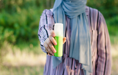 A Muslim woman in a blue hijab holds a bottle of mosquito repellent.