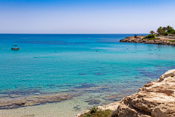 View of the Mediterranean Sea with clear water. The rocky coast of the resort village of Protaras on the island of Cyprus. Swimming people in the distance.