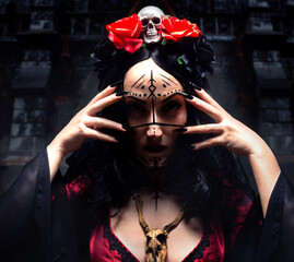 Beautiful mysterious sorceress or witch with runic makeup, wooden animal skull amulet and crown with skull and roses, touching her face at church background. Halloween concept.