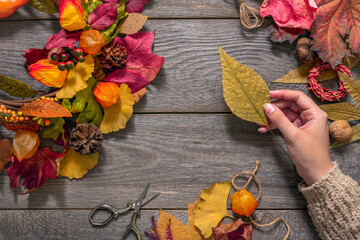 the process of creating autumn decorations, a female hand with a yellow dry leaf on a wooden background makes an autumn wreath