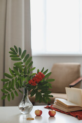 autumn still life with a rowan branch in a vase, book and apples in a cozy home interior