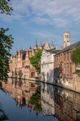 Brugge, Flanders, Belgium - August 4, 2021: Portrait, Belfry towers over the sunlighted facades of quiet Groenerei canal water mirroring the houses. Some green foliage on side.