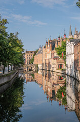 Brugge, Flanders, Belgium - August 4, 2021: Blinde Ezelstraat brick bridge and sunlighted palace building facades reflected in quiet Groenerei canal water under blue sky.