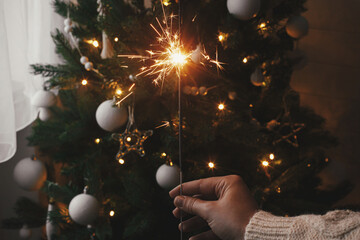 Happy New Year! Hand holding burning sparkler on background of christmas tree lights in festive...