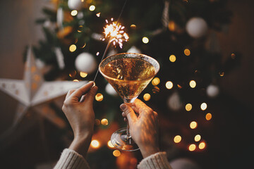 Happy New Year! Hands holding burning sparkler and champagne glass on background of christmas tree...
