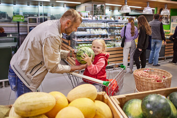 Happy little girl in shopping cart with father holds watermelon.