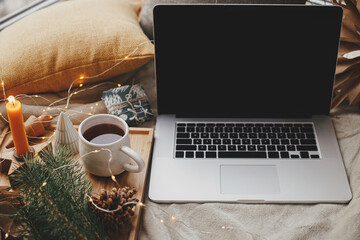 Laptop on soft bed with cup of tea, christmas lights, stars, pine, candle and pillows in...