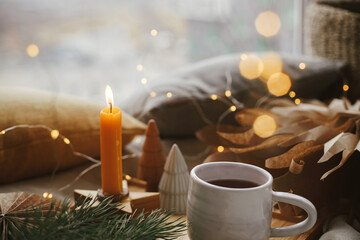 Warm cup of tea with christmas lights, stars, pine trees, candle and pillows on soft bed in...