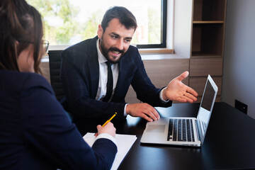 Handsome young office worker gesturing with hand, showing laptop screen while talking to female colleague.