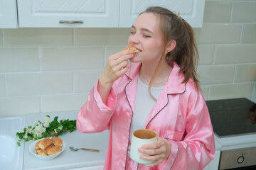beautiful young girl in pink silk pajamas has breakfast in the kitchen and happily eats a...
