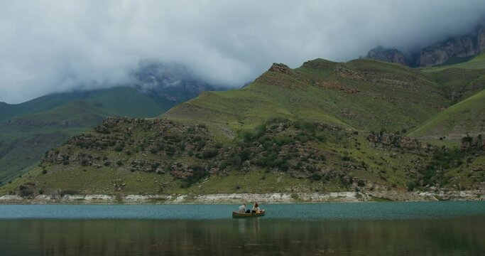 A couple are sitting in a rowboat in the lake against a backdrop of mountains.