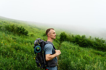 Hiker with big traveling rucksack moving up on the mountain trail among the tall grass.