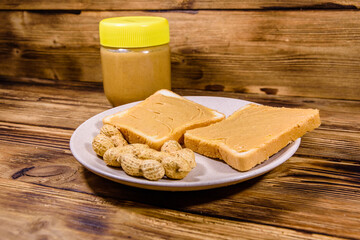 Glass jar with peanut butter and plate with sandwiches on a wooden table