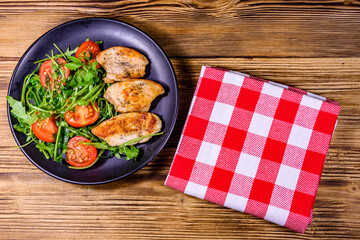 Roasted chicken breasts and salad with arugula and cherry tomatoes in a black plate. Top view