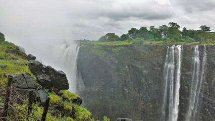 Fototapeta premium Streams of water flow down from the edge of the cliff. Everything is in a fog around. There is green vegetation on the plateau. Wet boulders in the foreground. Victoria Falls. Zimbabwe.