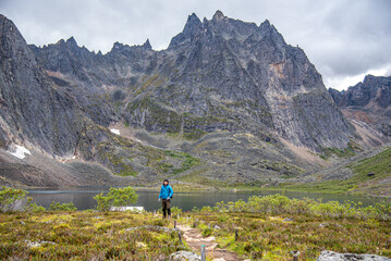 Man in blue jacket hiking through Tombstone Territorial Park during summer with towering mountain peaks behind. 